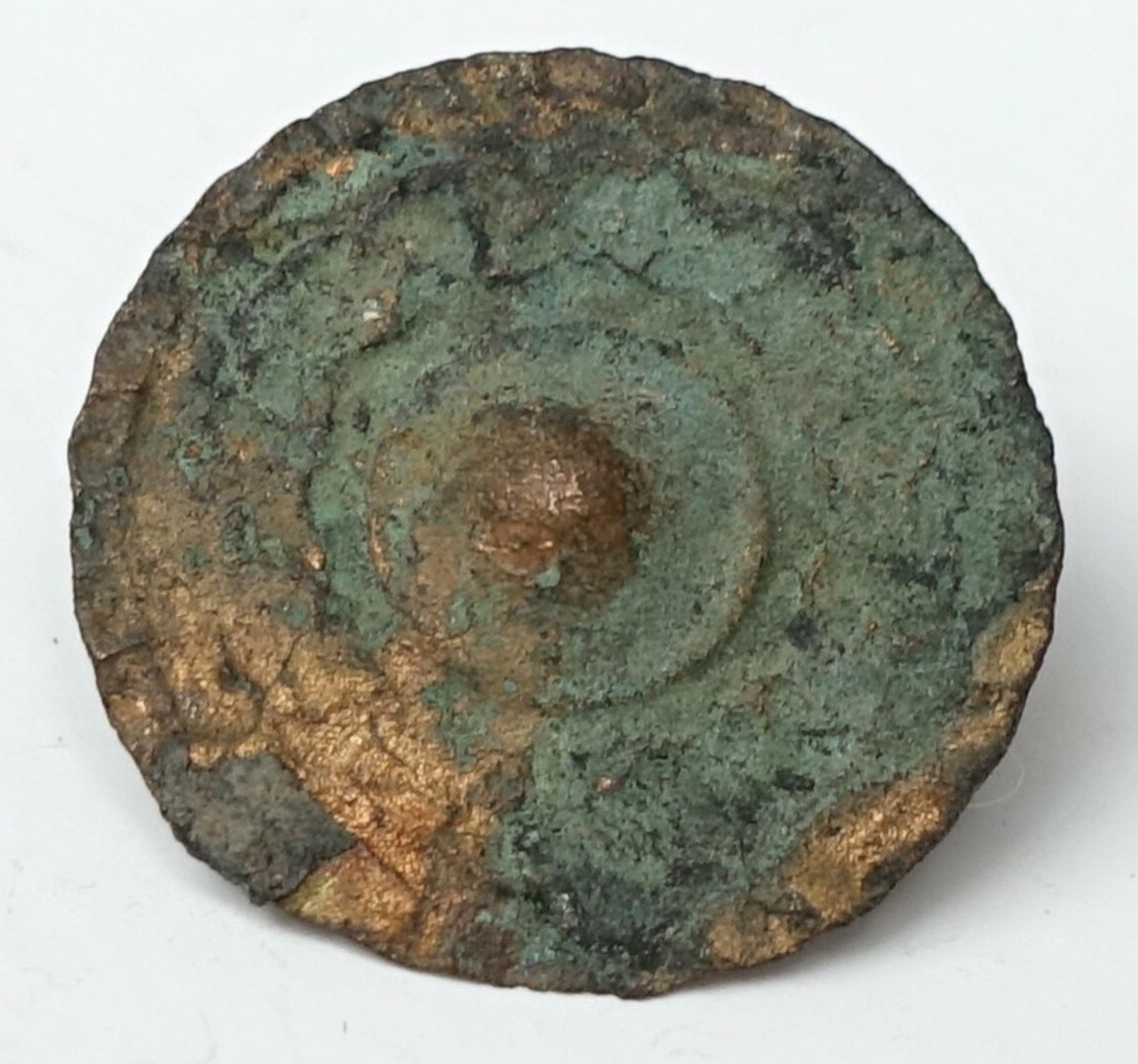 A metal button made of a thin, flat sheet with a domed centre and border decoration, with a small cone shank. The Netherlands, 19th century (TRC 2024.2592).