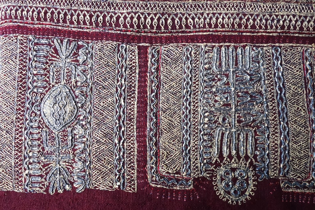 Fig. 1. Long, dark red cloth, decoratively woven with narrow mid-red stripes at regular intervals. Across the middle of the cloth there is a broad band of silver thread embroidery with stripes of repeating, geometric motifs. Tunisia, first half 20th century (TRC 2026.0187).