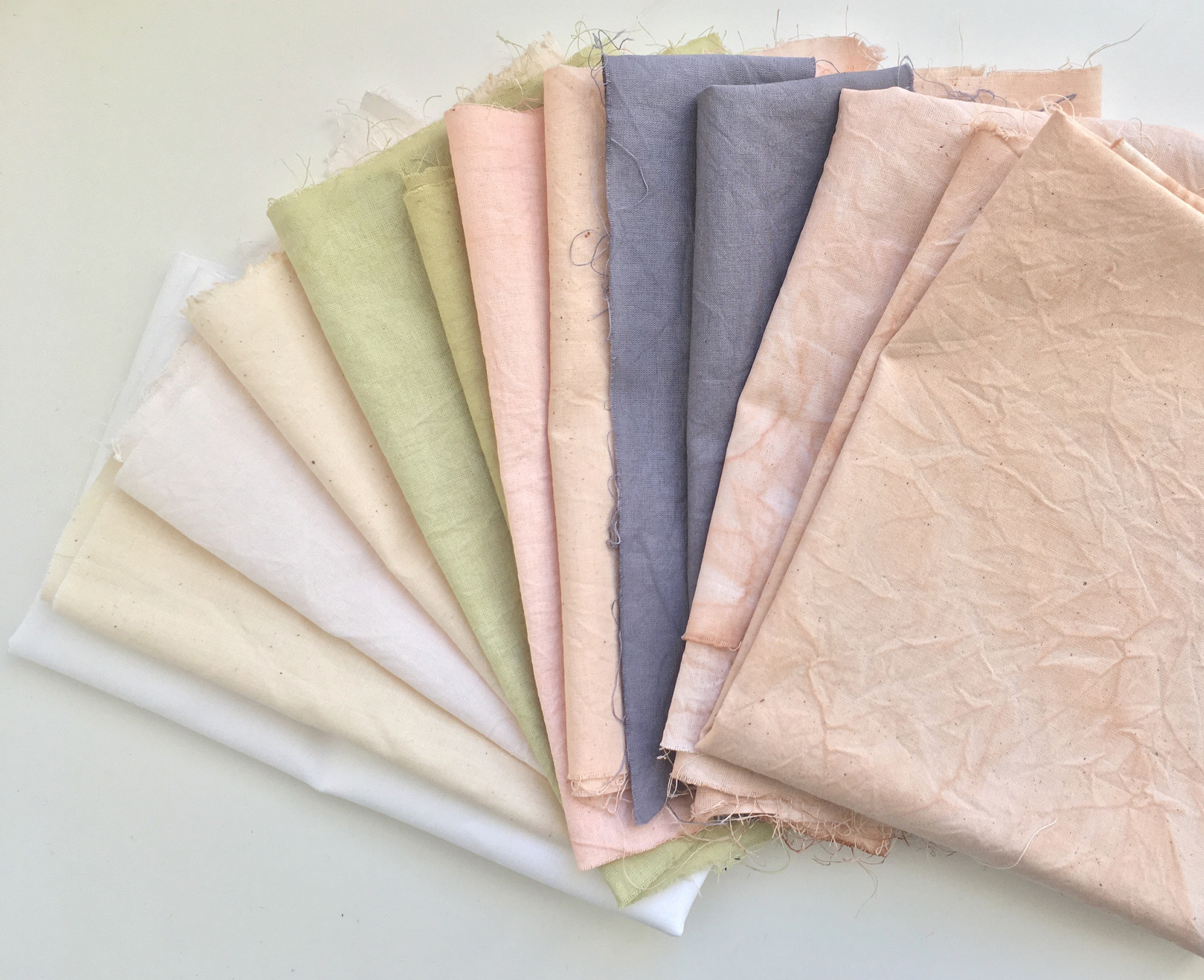 Natural and white cotton fabrics dyed with kitchen waste. From left to right: Original fabric colour, mango peels, nettles, Avocado skin and pits, black beans, cassava.